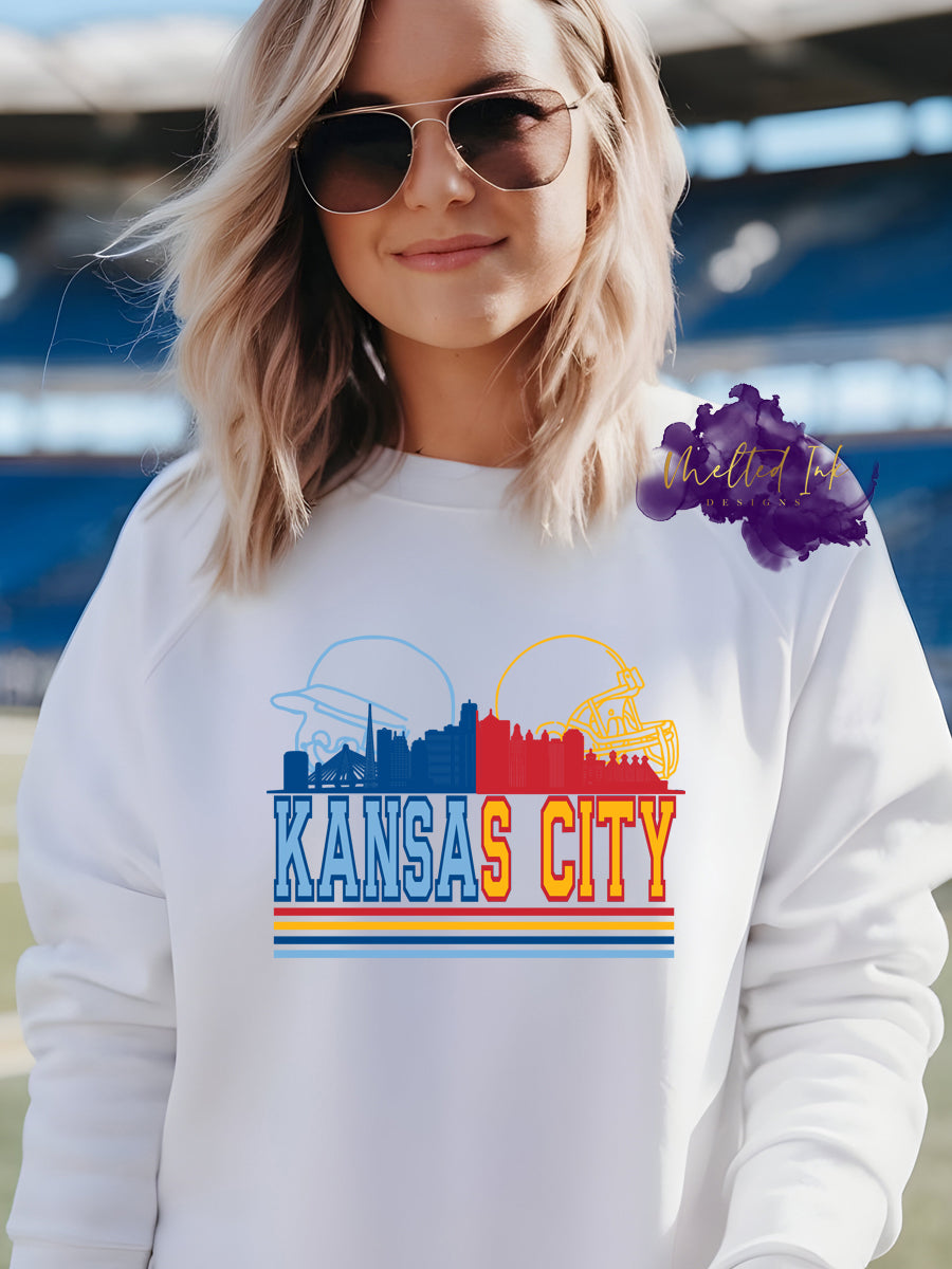 Photo is a mock up of design and is a picture of the Kansas City Skyline. Half of it is blue and half of it is red. Behind the blue part of the skyline is a baseball helmet and behind the red side is a yellow outline of a football Helmet. Below the skyline is the words Kansas City and those are also blue and yellow.