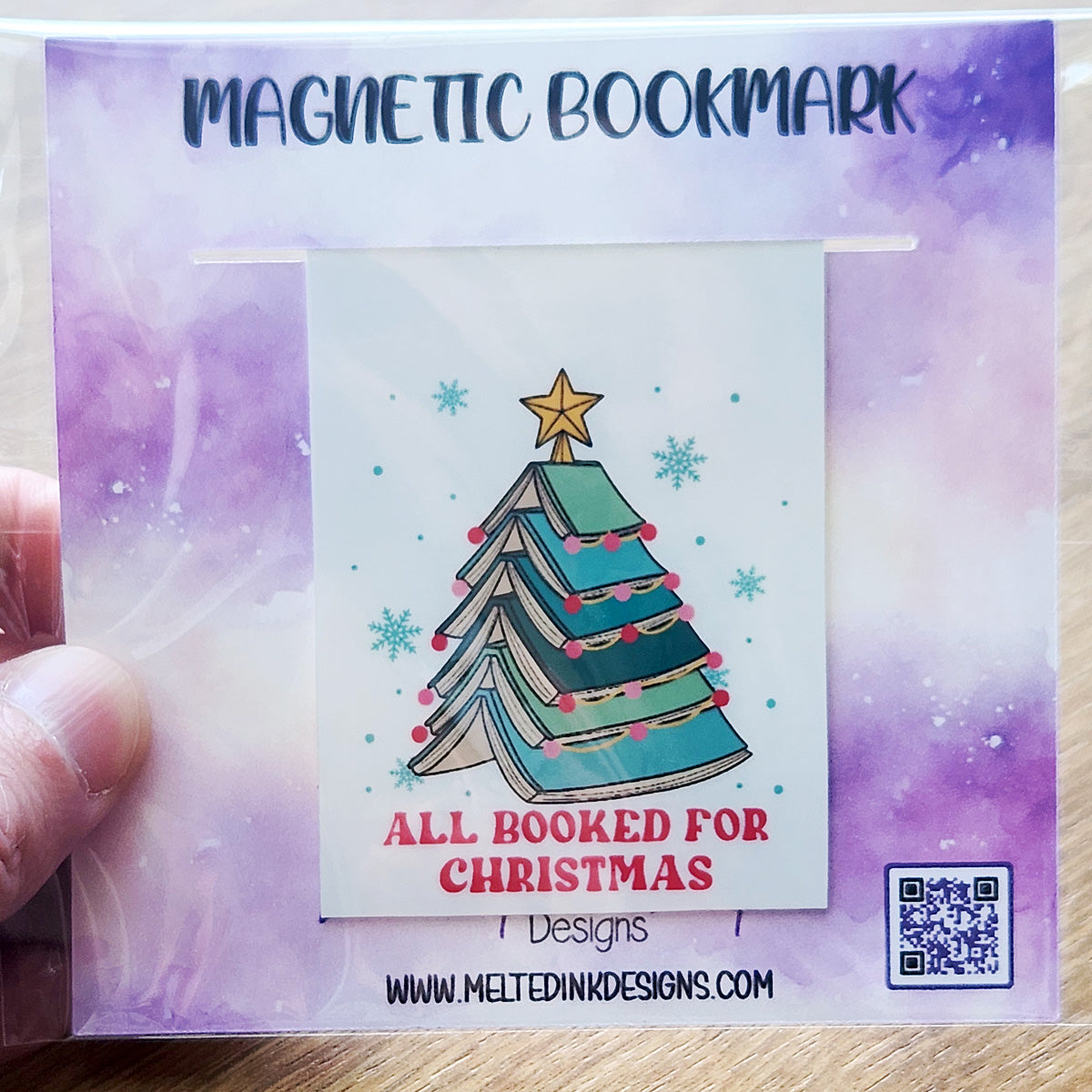 This magnetic bookmark features a festive design of a stack of opened books arranged in the shape of a Christmas tree, topped with a star and features the phrase "All Booked for Christmas"
