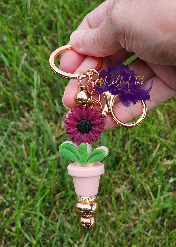 photo is a rose gold keychain beadable bar that has a burgandy sunflower with a pink flower pot underneath and is accented with a rhintestone spacer
