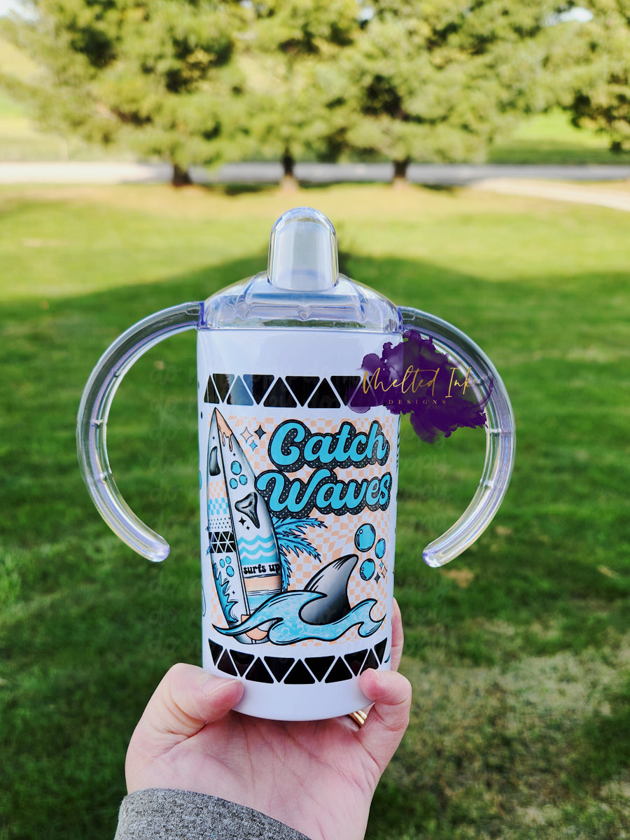 Photo consists of a kids sippy cup that converts into a tumbler. The design on the cup consists of a surfboard with a shark in the wave next to it and the cup says Catch Waves.