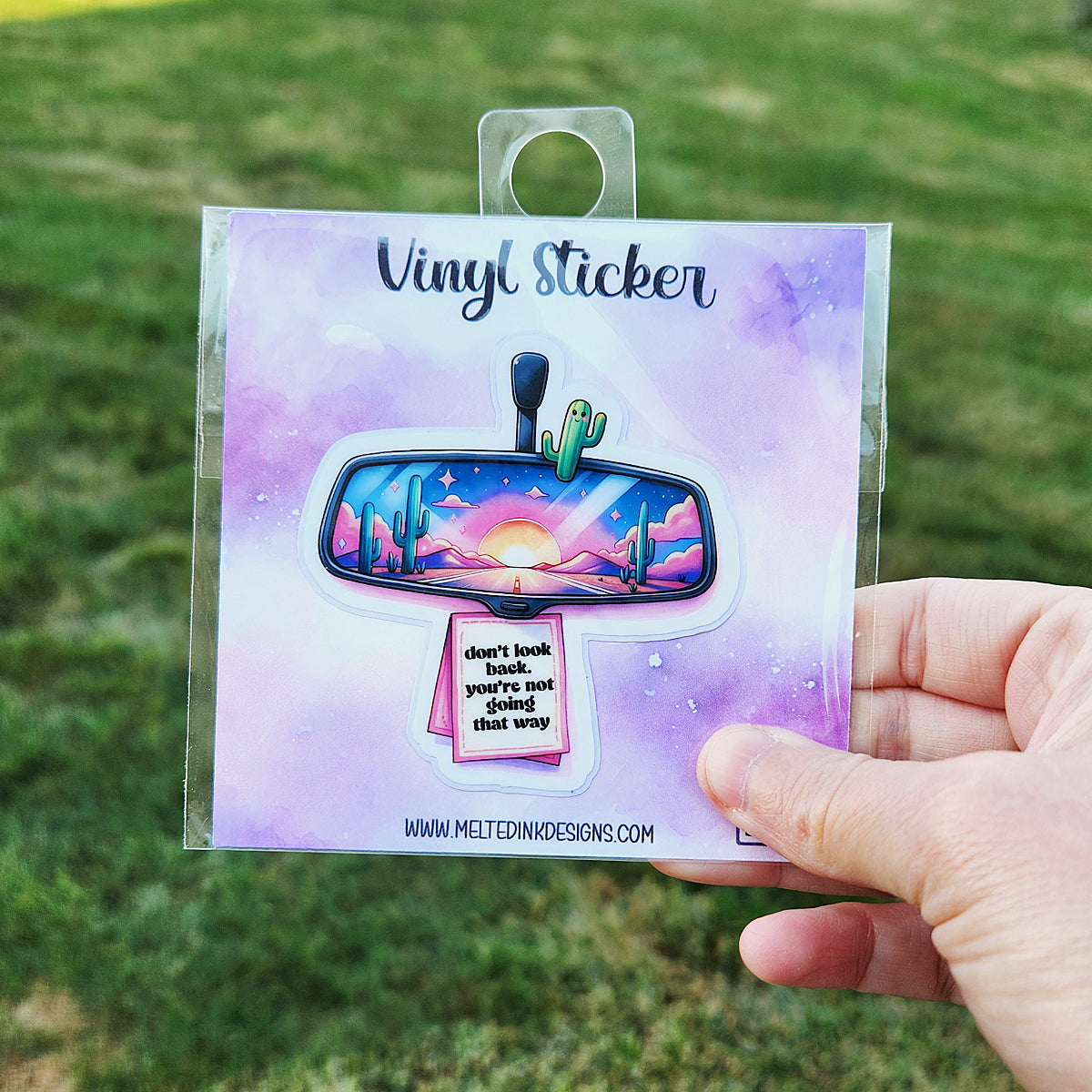 Photo is of a sticker that has a rearview mirror in it. In the review mirror there is a sunset with cactus and it says Don't look back your not going that way.