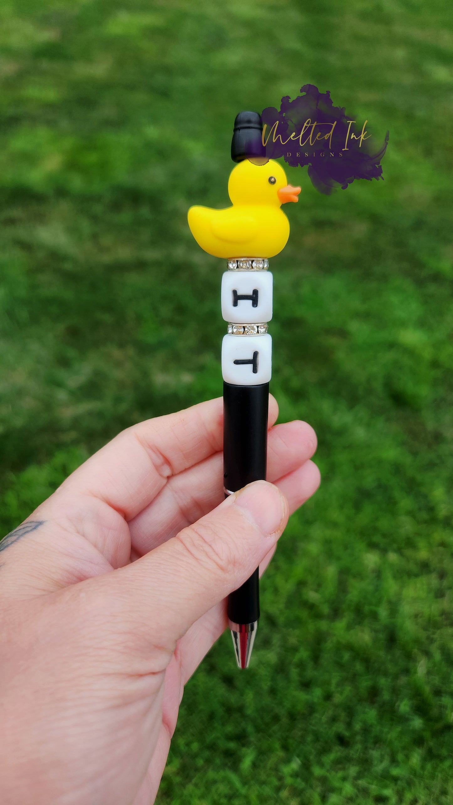 Photo is beaded black ink pen that showcases a yellow duck focal bead. Underneath the duck are letter silicone beads that say IT with spacers inbetween. Ink is black.