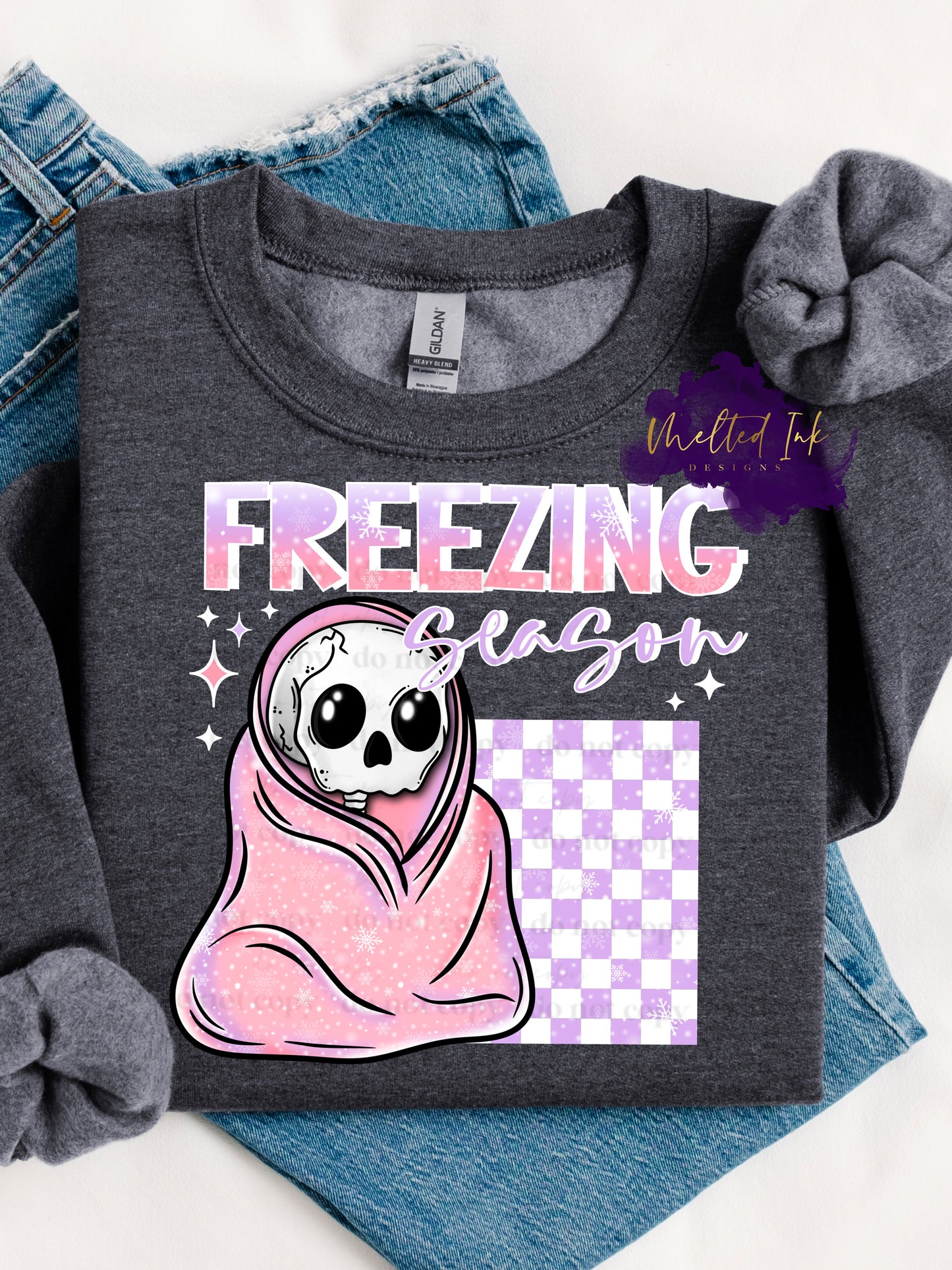 Design is a mock up and is on a dark gray shirt. Design is pink/purple in color and says Freezing Season. Has a skeleton wrapped up in a blanket.