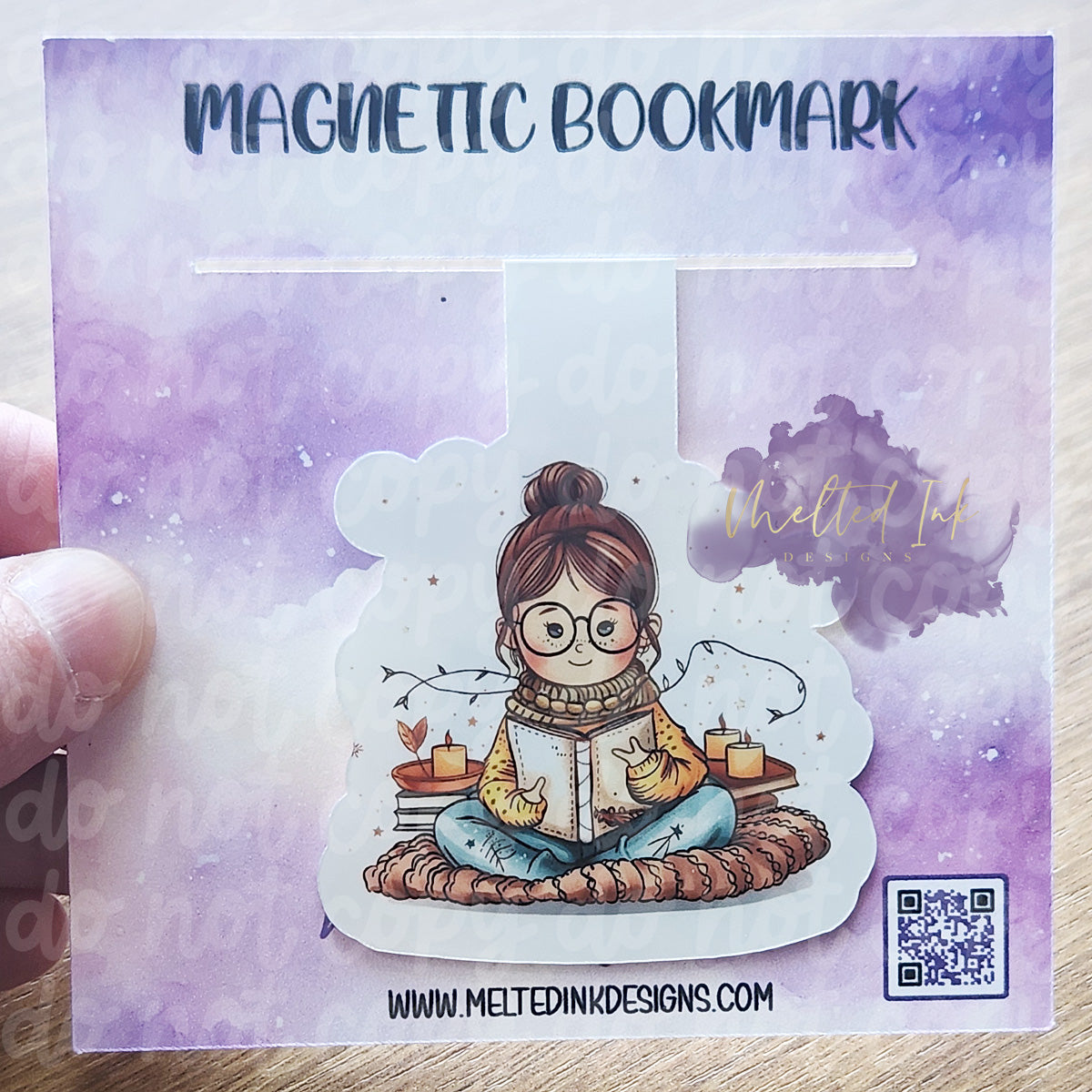 Photo is of a magnetic bookmark. Shows a girl sitting on a blanket on the floor reading a book. In the background it has some stacked books with candles sitting on top.