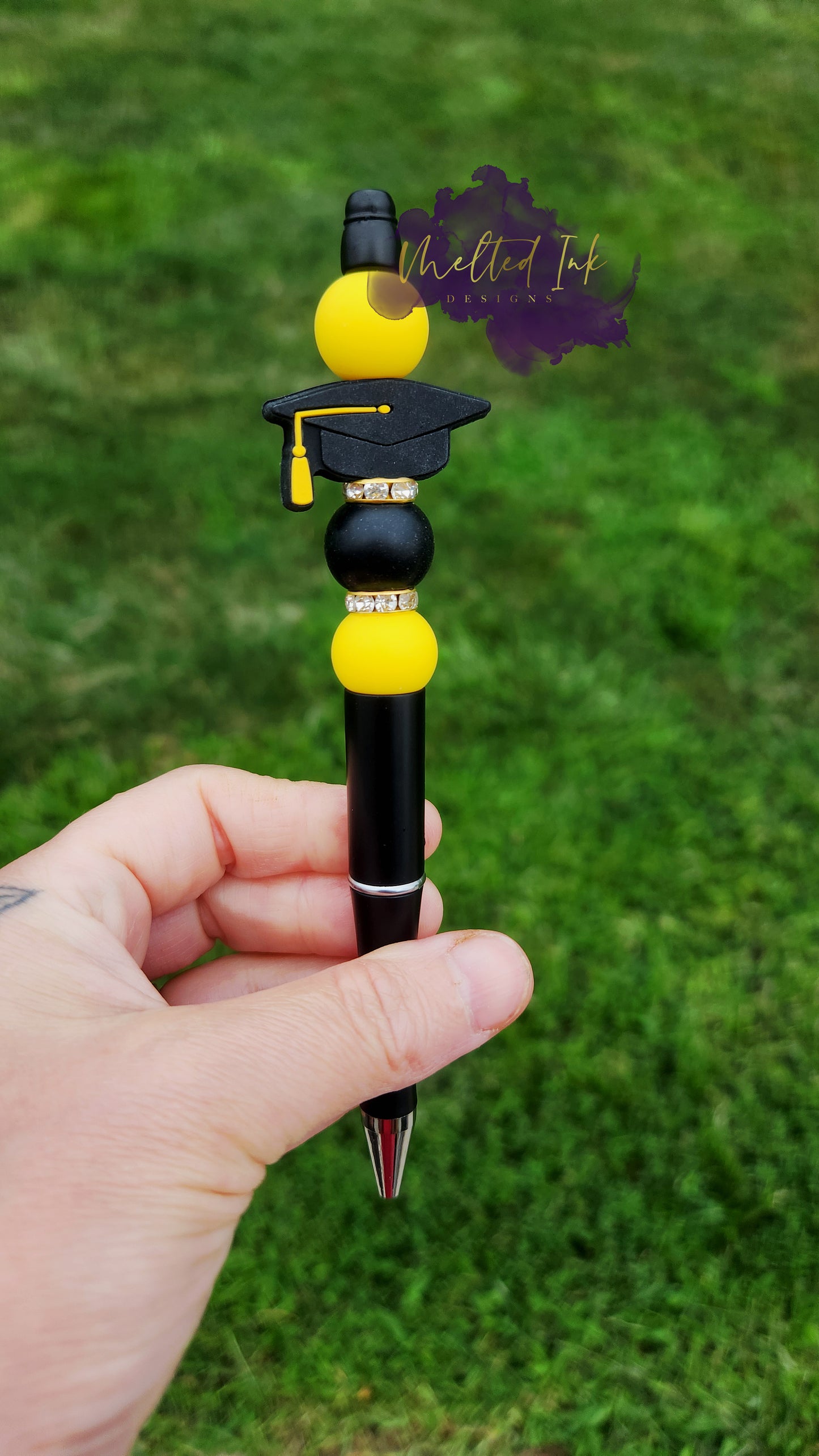 Photo is beaded black ink pen that showcases a black with yellow tassle graduation cap focal bead. Surrounding the focal bead is black and yellow silicone beads with rhinestone spacers. Ink is black.