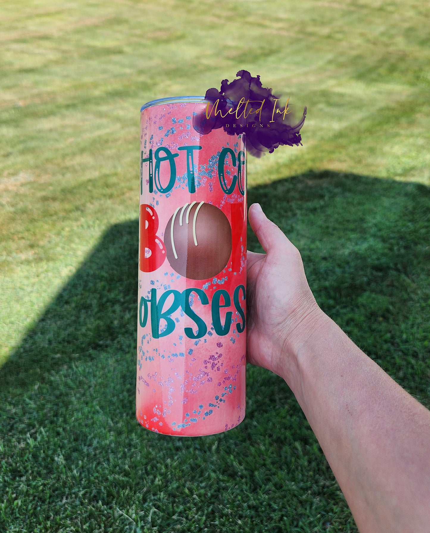 Photo consists of a 20oz tumbler that says hot cocoa bomb with the O in bomb being a chocolate covered cocoa bomb. Background is a pinkish red color.