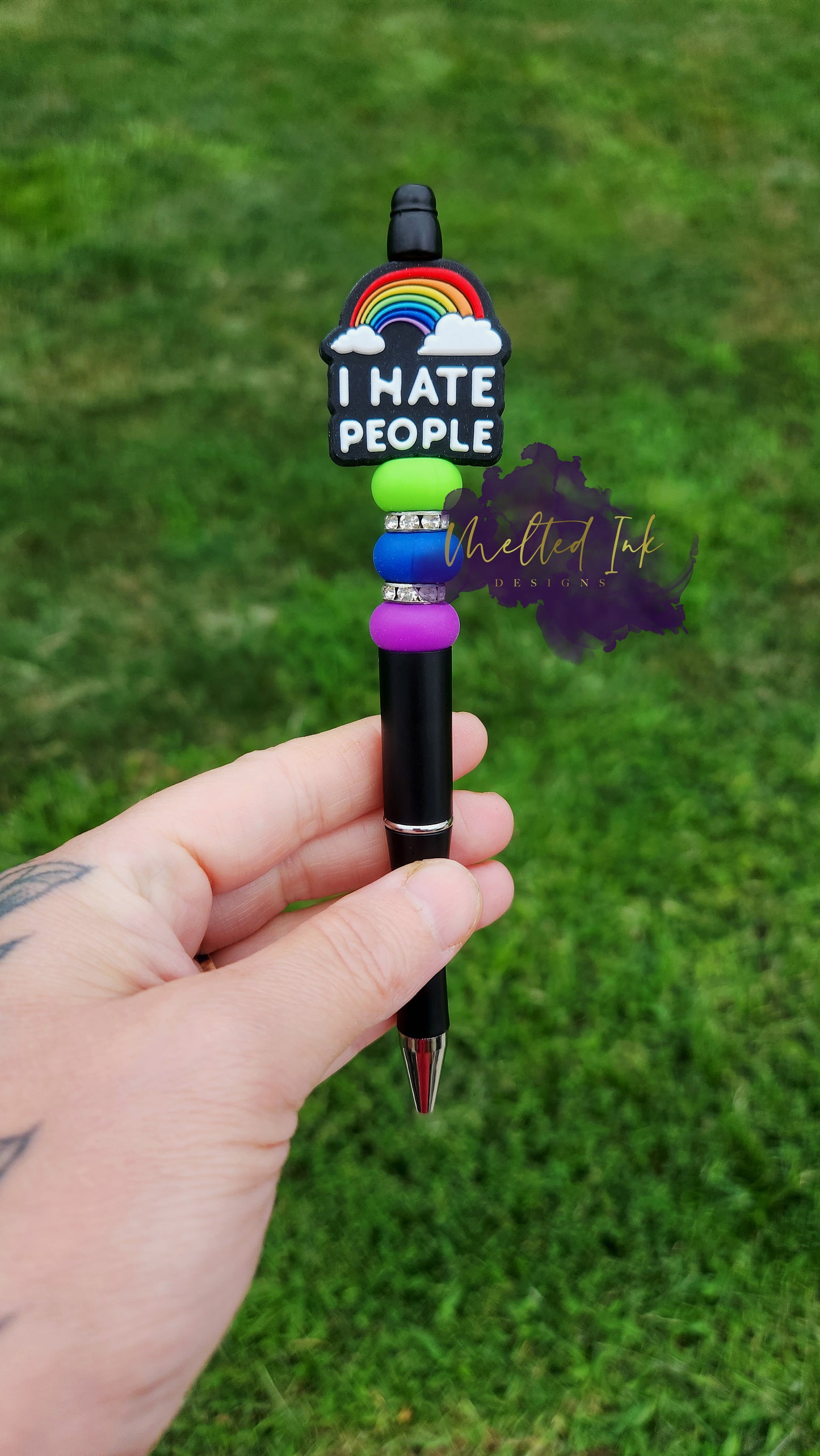 Photo is a black beaded ink pen that has a focal with a rainbow that says I hate people. It is accented with a lime green, blue and purple silicone spacer bead and also has rhinestone spacers. Ink is black.