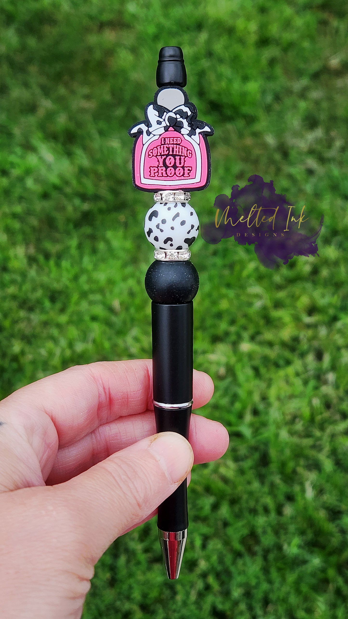 Photo is a black beaded ink pen that has an alcohol bottle focal bead that says I need something you proof and has a cow print bow on it. Accenting the focal bead is a black cow print and a black with white speckled silicone beads and rhinestone spacers. Ink is black.