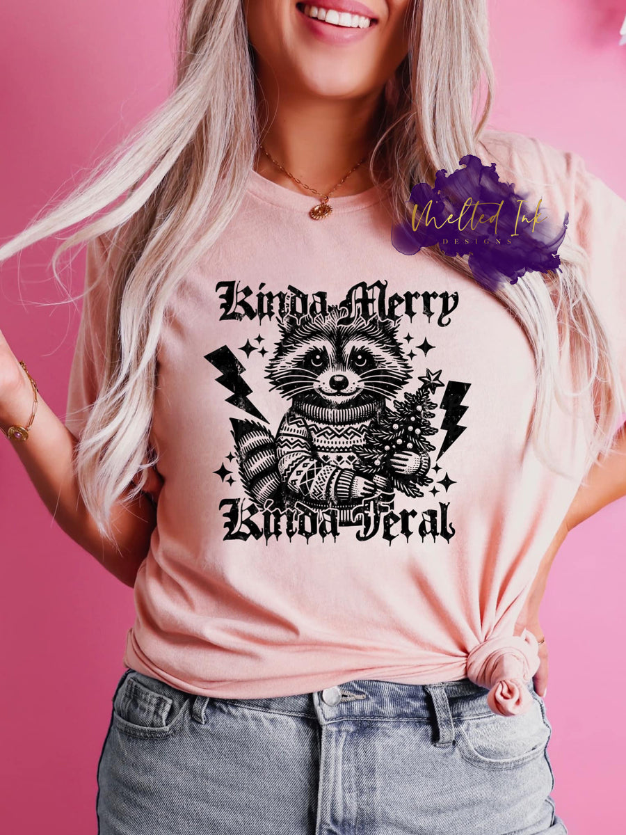 Picture is a mock up of design. Design is black in color and says Kinda Merry Kinda Feral and has a raccoon with a Christmas sweater holding a Christmas tree.