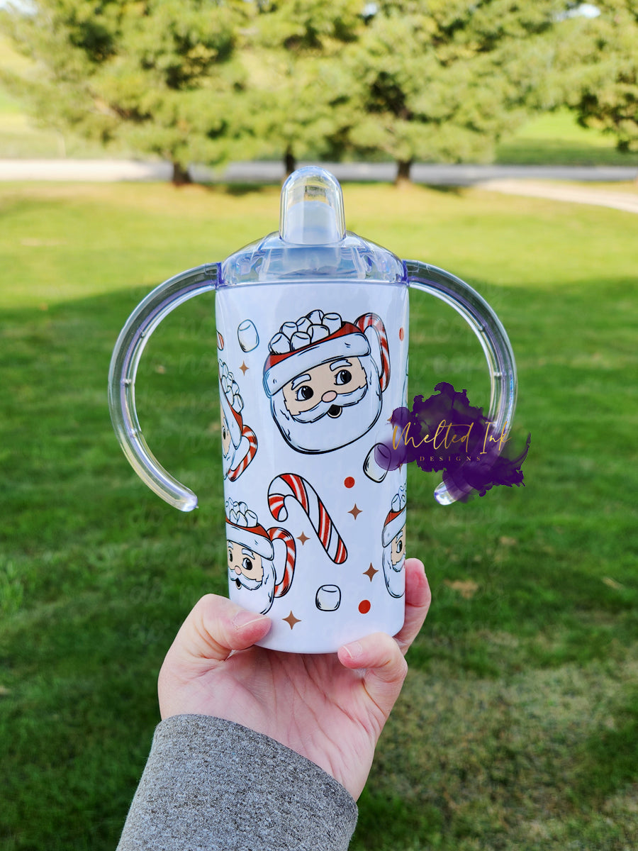 Photo consists of a kids sippy cup that converts into a tumbler. The design on the cup consists of santa face mugs filled with marshmellows. Background has marshmellows, candy canes, and little dots and stars.