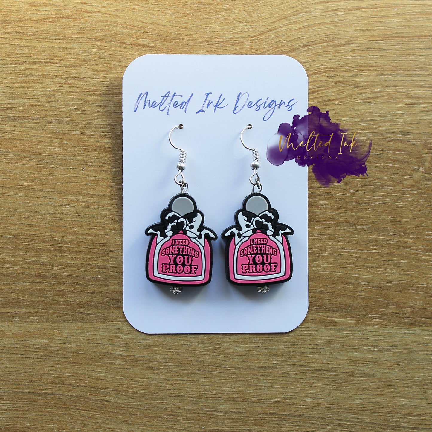 Photo is a picture of a pair of little pink alcohol bottle earrings that says I need something you proof. Also has a cow print bow on each earring.