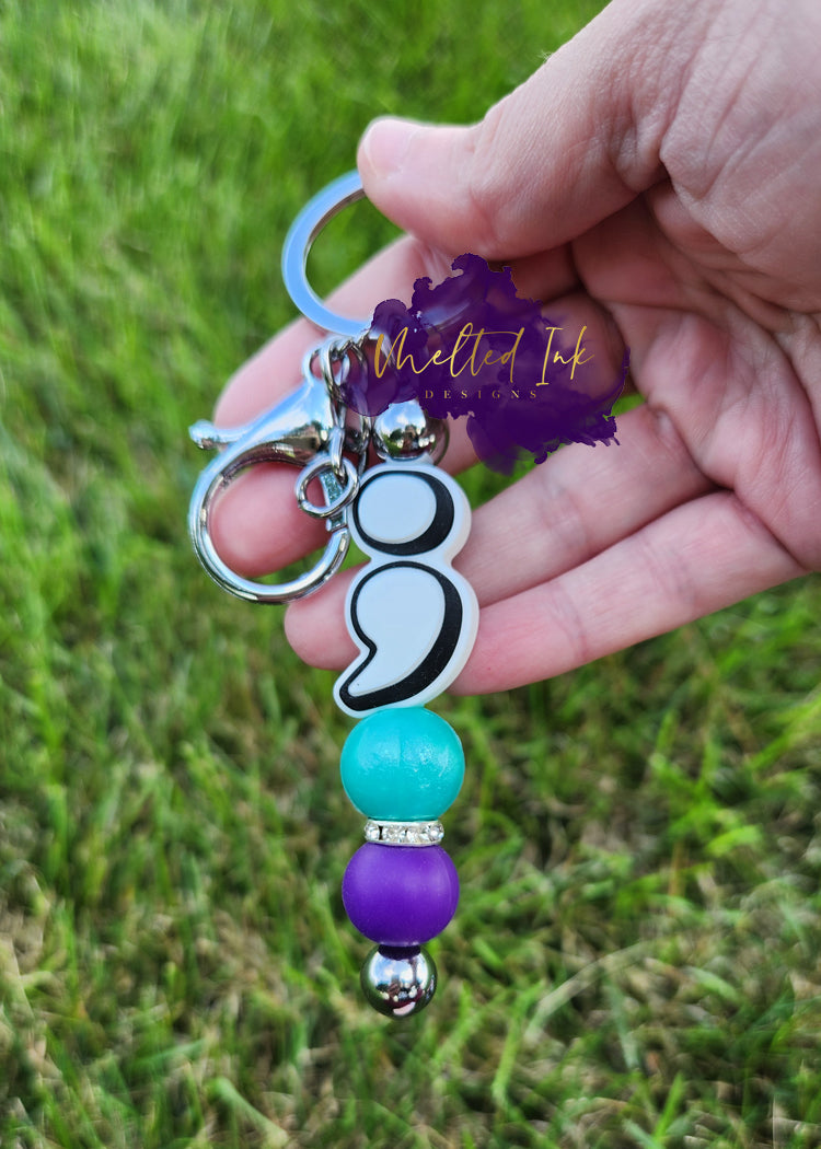 he photo showcases a silver beadable keychain bar designed with a meaningful message for suicide awareness. At the top, there’s a semi-colon focal bead, with the opposite side saying the phrase "It's okay to not be okay". The keychain is adorned with light blue and purple solid silicone beads, and has a rhinestone accent adds a bit of sparkle, enhancing the overall design.