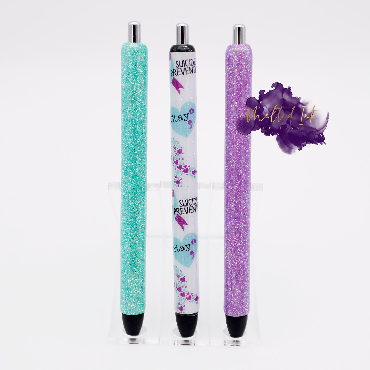Photo is a set of 3 epoxy resin ink pens for suicide prevention. It has one solid tiffany blue and a light purple glitter ink pen. The other pen included has a pen wrap that has suicide prevention and the word stay in a heart with a semicolon. This one also includes confetti type hears in the shape of a suicide awareness ribbon. The ink is black.