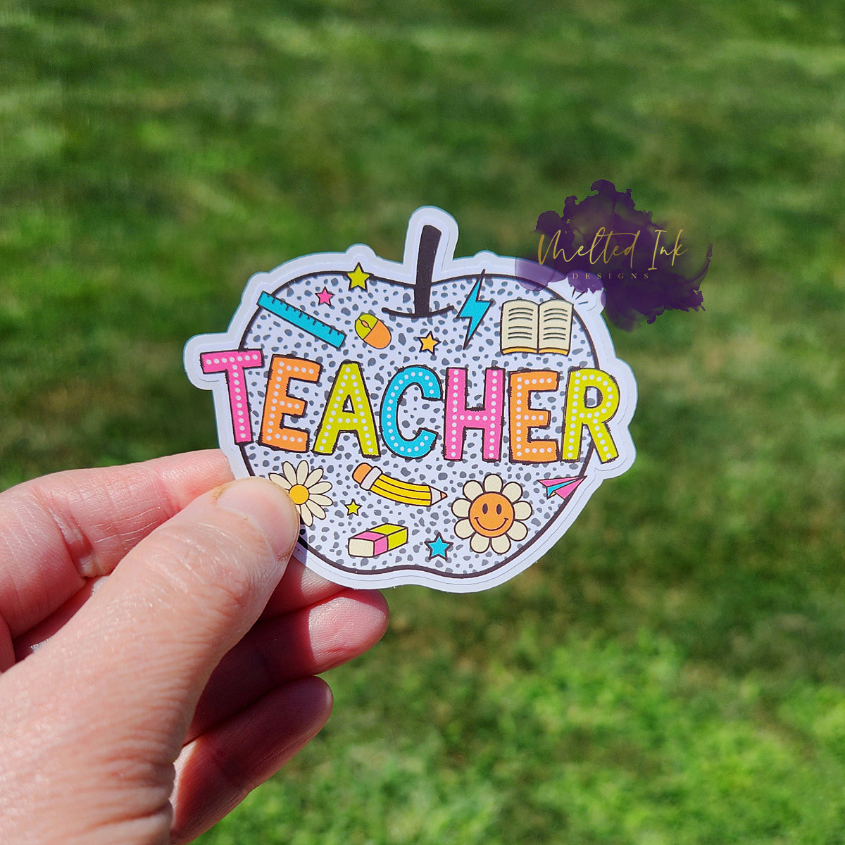 Photo is of a sticker that says teacher inside of an apple that is white. The apple has gray polka dots and has different little teacher elements such as a ruler, book, pencil, eraser, flowers, etc.
