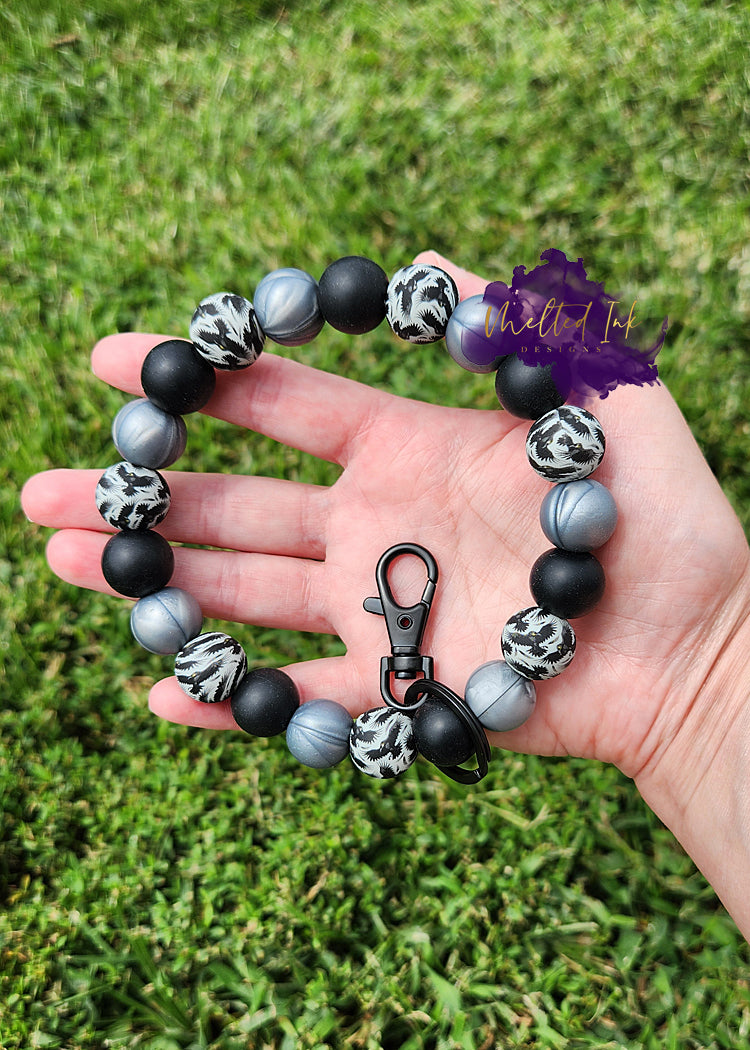 This stylish wristlet features alternating silicone beads adorned with a striking black raven print, paired with elegant black and grey marble beads. Comes with keyring.