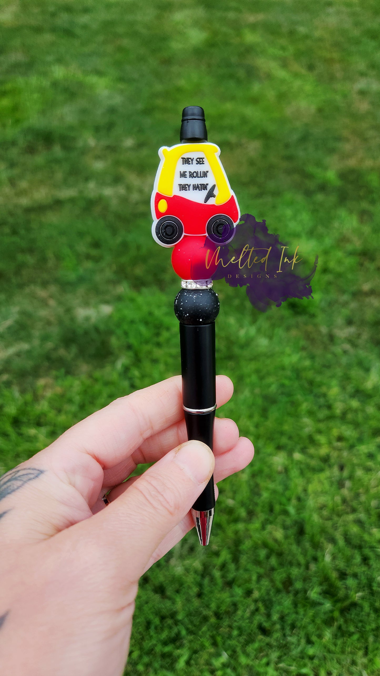 Photo is a little toy tykes car that says they see me rolling they hatin. Accenting the focal bead is a solid red and a black with white splatter silicone bead and includes a rhinestone spacer inbetween. Ink is black.