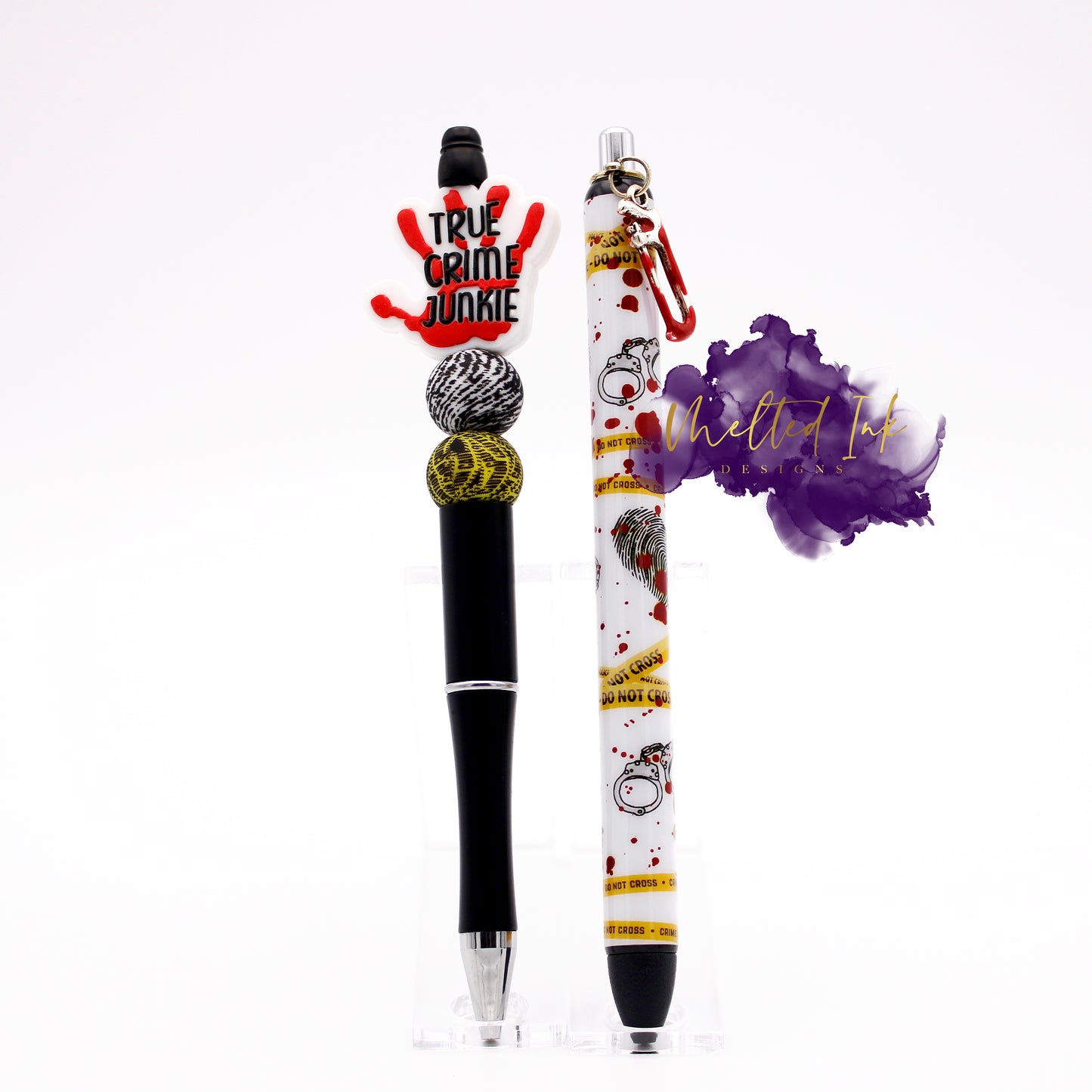 This photo consists of a true crime ink pen set. It has a beaded black ink pen that has a bloody hand print focal bead that says true crime junkie with a finger print and caution take silicone beads. Also included is an epoxy pen that has caution tape, handcuffs and finger print with blood splatters. It has a little saw charm with fake blood at the top. Both ink pens have black ink.
