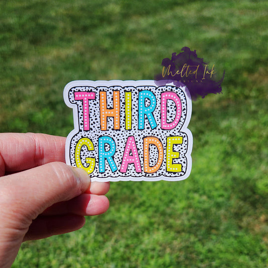 Photo is of a sticker that says third grade and has a black polka dot outline around it. 