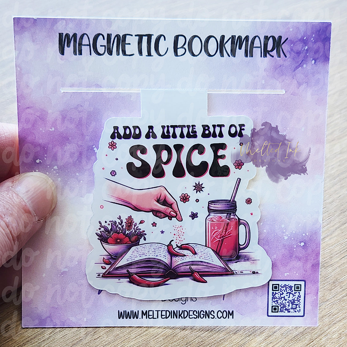 This magnetic bookmark has a fun and flavorful theme with the phrase "Add a little bit of spice." It features a hand sprinkling spice onto an open book, complemented by little jalapeños placed on the pages. The design also includes a charming bowl of flowers and a refreshing drink in a mason jar.