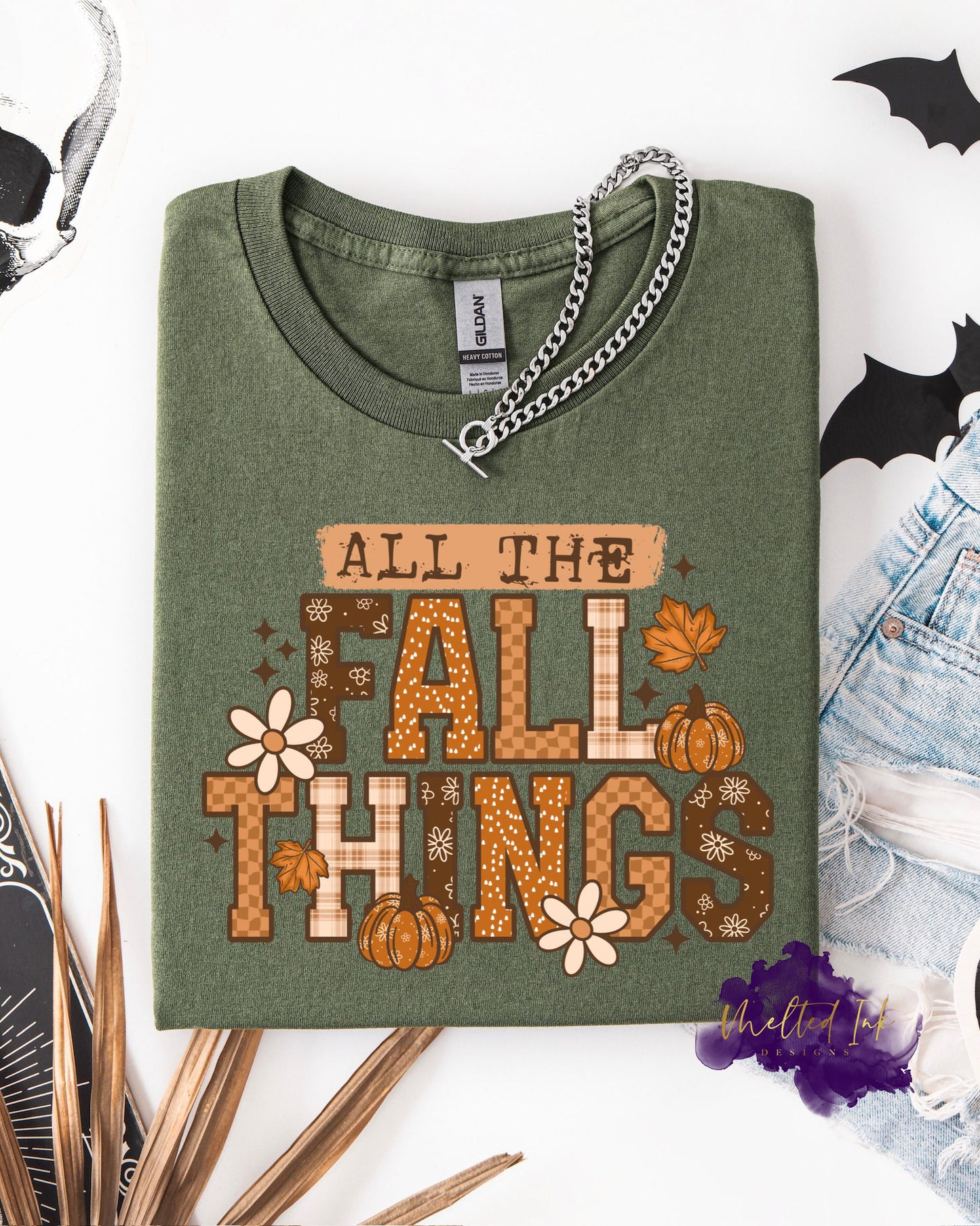 A shirt with the words “ALL THE FALL THINGS” in bold lettering with plaid textures, pumpkins, flowers, and fall leaves. A cozy autumn design perfect for fall shirts, sweatshirts, and seasonal apparel.