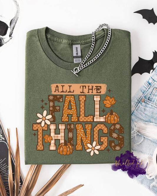 A shirt with the words “ALL THE FALL THINGS” in bold lettering with plaid textures, pumpkins, flowers, and fall leaves. A cozy autumn design perfect for fall shirts, sweatshirts, and seasonal apparel.