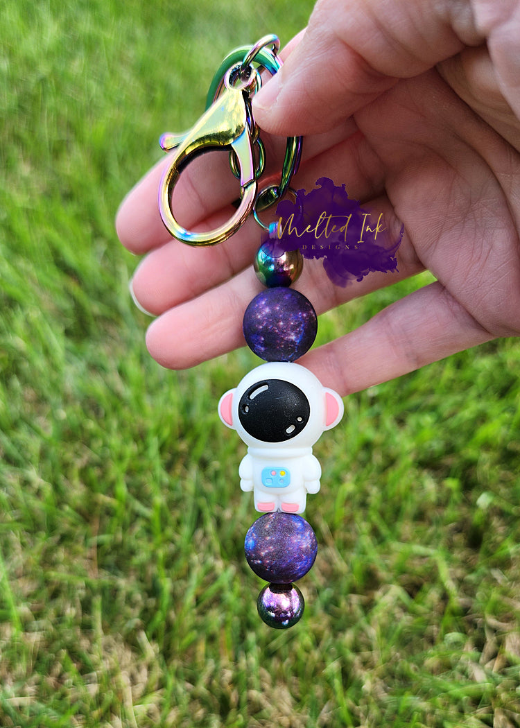 The photo features a beadable keychain bar that prominently displays an astronaut focal bead in the center. Above and below the astronaut, there are galaxy focal beads, likely adorned with vivid colors and cosmic designs that enhance the space theme.