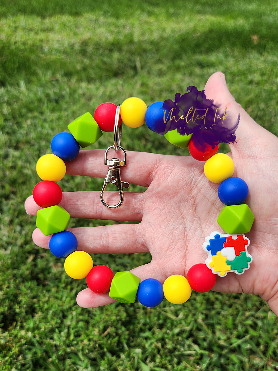 Photo is an autism awareness wristlet in the colors of red, blue, yellow and green. It has a focal bead that consists of the autism puzzle and includes a silver keyring. 