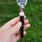 Photo is beaded black ink pen that showcases a round focal bead that says best grandma ever. Accenting the focal bead is a pink floral printed and a light pink silicone bead. A rhinestone spacer has been added to add a bit of sparkle. Ink is black. 