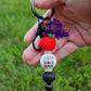 photo showcases a black beadable keychain bar featuring an apple focal bead at the top symbolizing education. Below the apple, there’s an acrylic bead that reads "Coffee, Teach, Repeat," which adds a fun and relatable touch for teachers. Complementing these focal beads is a solid black silicone bead on the bottom.