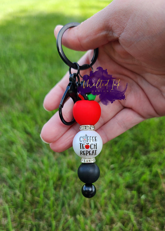 photo showcases a black beadable keychain bar featuring an apple focal bead at the top symbolizing education. Below the apple, there’s an acrylic bead that reads "Coffee, Teach, Repeat," which adds a fun and relatable touch for teachers. Complementing these focal beads is a solid black silicone bead on the bottom.