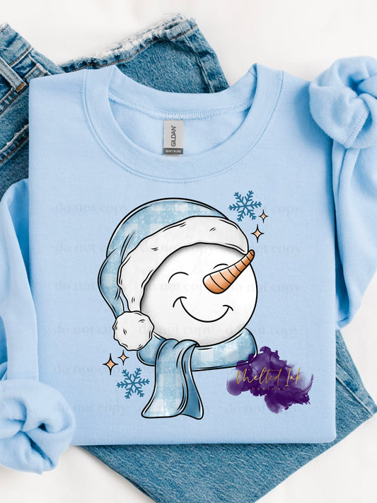 Picture is a mock up of design on a light blue sweatshirt. Picture is white and blue in color and is of a snowman wearing a plaid hat and scarf. 