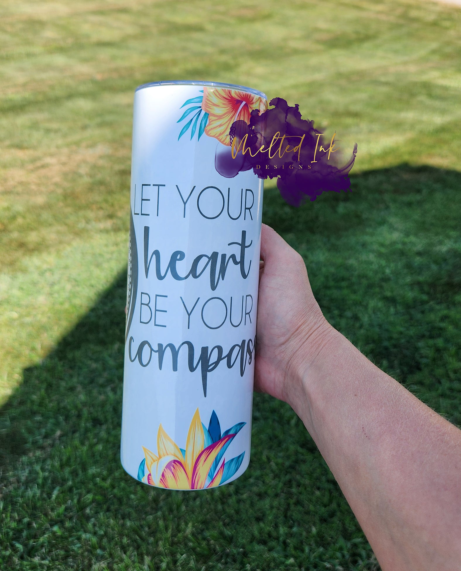 Photo features a 20oz tumbler that says let your heart be your compass. Shows a compass sign and has yellowish red lilly flowers. 