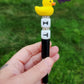 Photo is beaded black ink pen that showcases a yellow duck focal bead. Underneath the duck are letter silicone beads that say IT with spacers inbetween. Ink is black.  