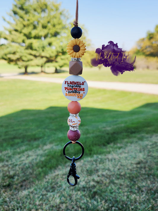 This fall lanyard features a cheerful yellow sunflower focal bead and another focal bead that lists "Flannels, Hayrides, Pumpkins, Sweaters, and Bonfires." The design is beautifully accented with silicone beads and rhinestones in warm autumn colors, creating a cozy and seasonal vibe. It also includes a keyring for added convenience.