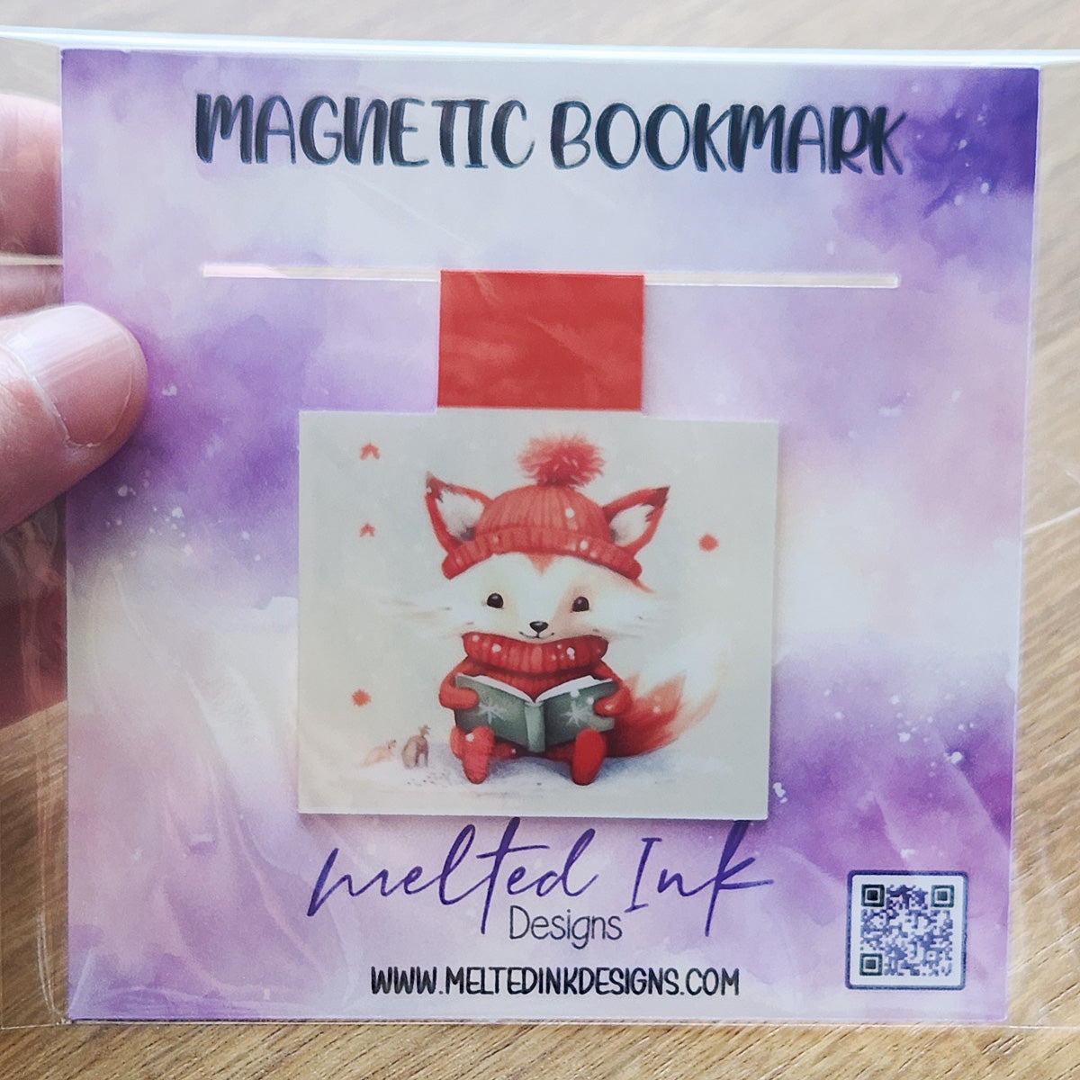 This magnetic bookmark features an adorable orange fox dressed in a cozy winter hat and sweater, happily reading a book. The charming illustration captures a warm and whimsical winter vibe.