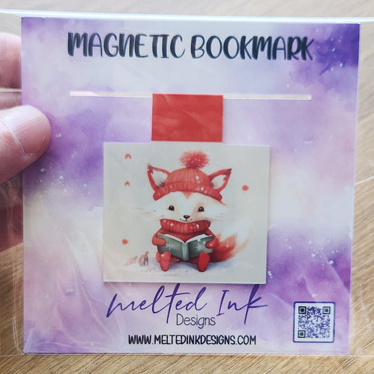 This magnetic bookmark features an adorable orange fox dressed in a cozy winter hat and sweater, happily reading a book. The charming illustration captures a warm and whimsical winter vibe.