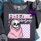 Design is a mock up and is on a dark gray shirt. Design is pink/purple in color and says Freezing Season. Has a skeleton wrapped up in a blanket. 