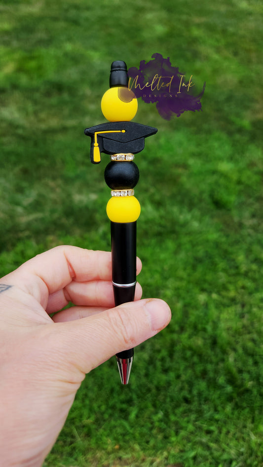 Photo is beaded black ink pen that showcases a black with yellow tassle graduation cap focal bead. Surrounding the focal bead is black and yellow silicone beads with rhinestone spacers. Ink is black. 