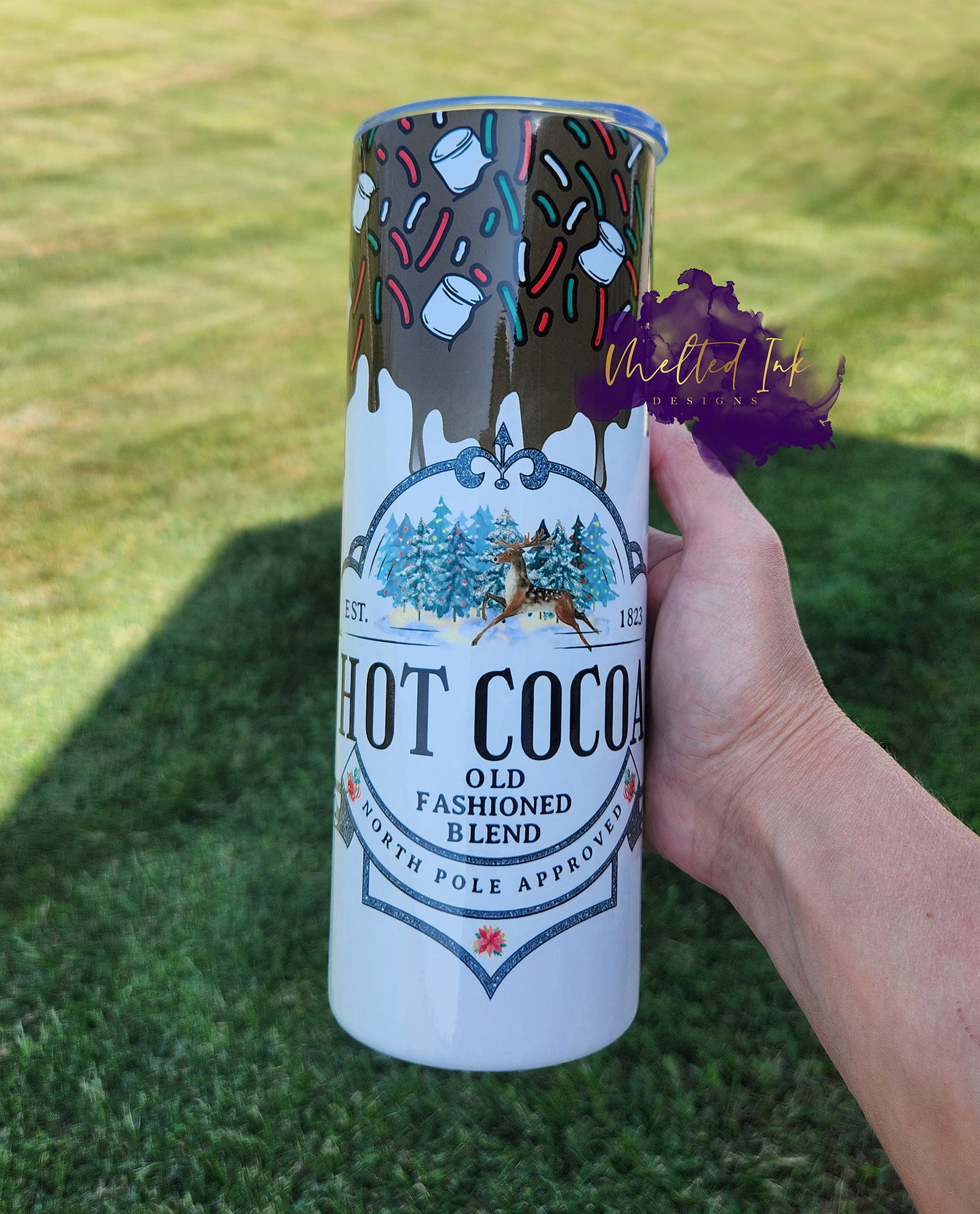 Photo is a 20oz tumbler and has melting chocolate at the top dripping down with marshmellows and red, green and white candy sprinkles. Cup says hot cocoa old fashioned blend north pole approved and has a picture of a deer in front of some christmas trees. 