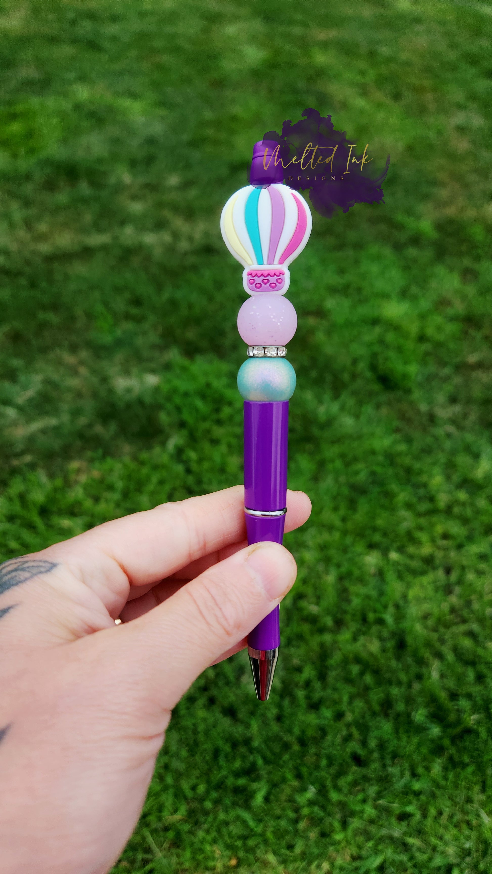 Photo is beaded purple ink pen that showcases a hot air balloon focal bead. Surrounding the focal bead are sparkling purple glitter and bluish-green pearl silicone beads, adding a touch of color and whimsy. The design is enhanced by a rhinestone spacer, which adds a bit of sparkle and elegance. Pen ink is black. 