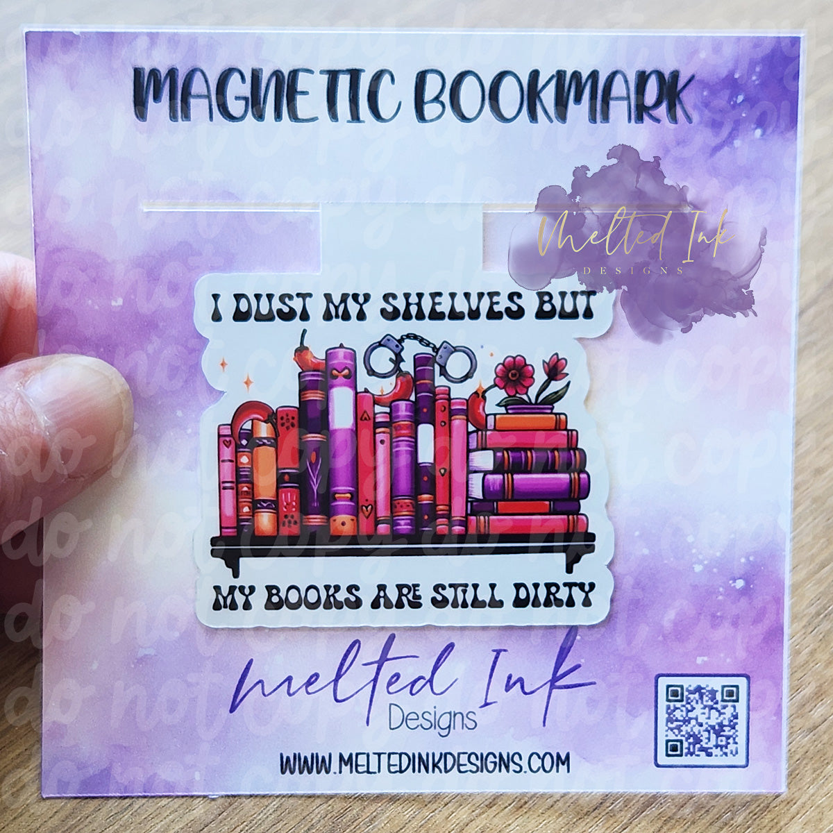 The bookmark features a whimsical design with the text, "I dust my shelves but my books are still dirty." Surrounding the phrase are colorful illustrations, including a stack of books, handcuffs, vibrant flowers, and jalapeños.