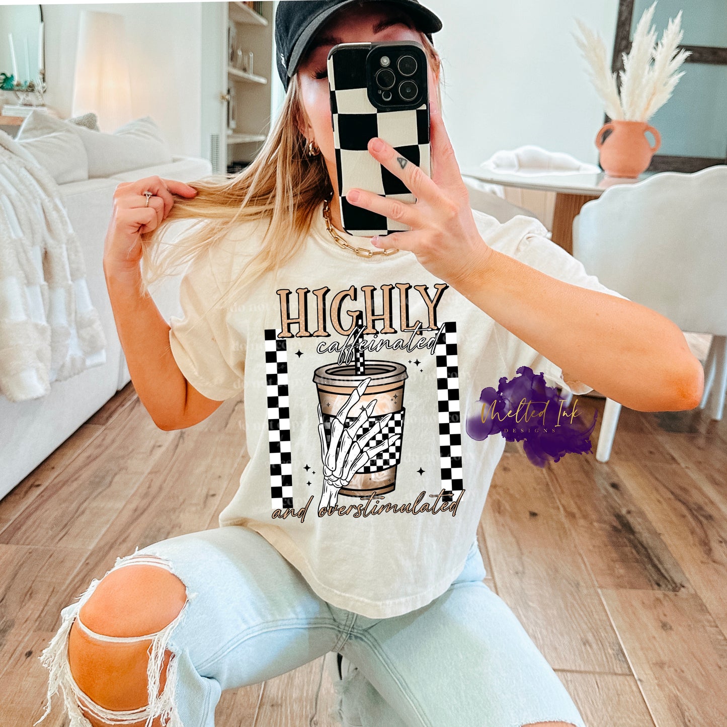 Photo is a mock up of the design. Design print is colored and pressed on an off white shirt. Design says Highly caffeinated and overstimulated. Picture of design has a skeleton hand holding a coffee cup with a straw. Cup wrap is black and white checkered. Design also has a checkered frame going vertical on the left and right side of print design. 