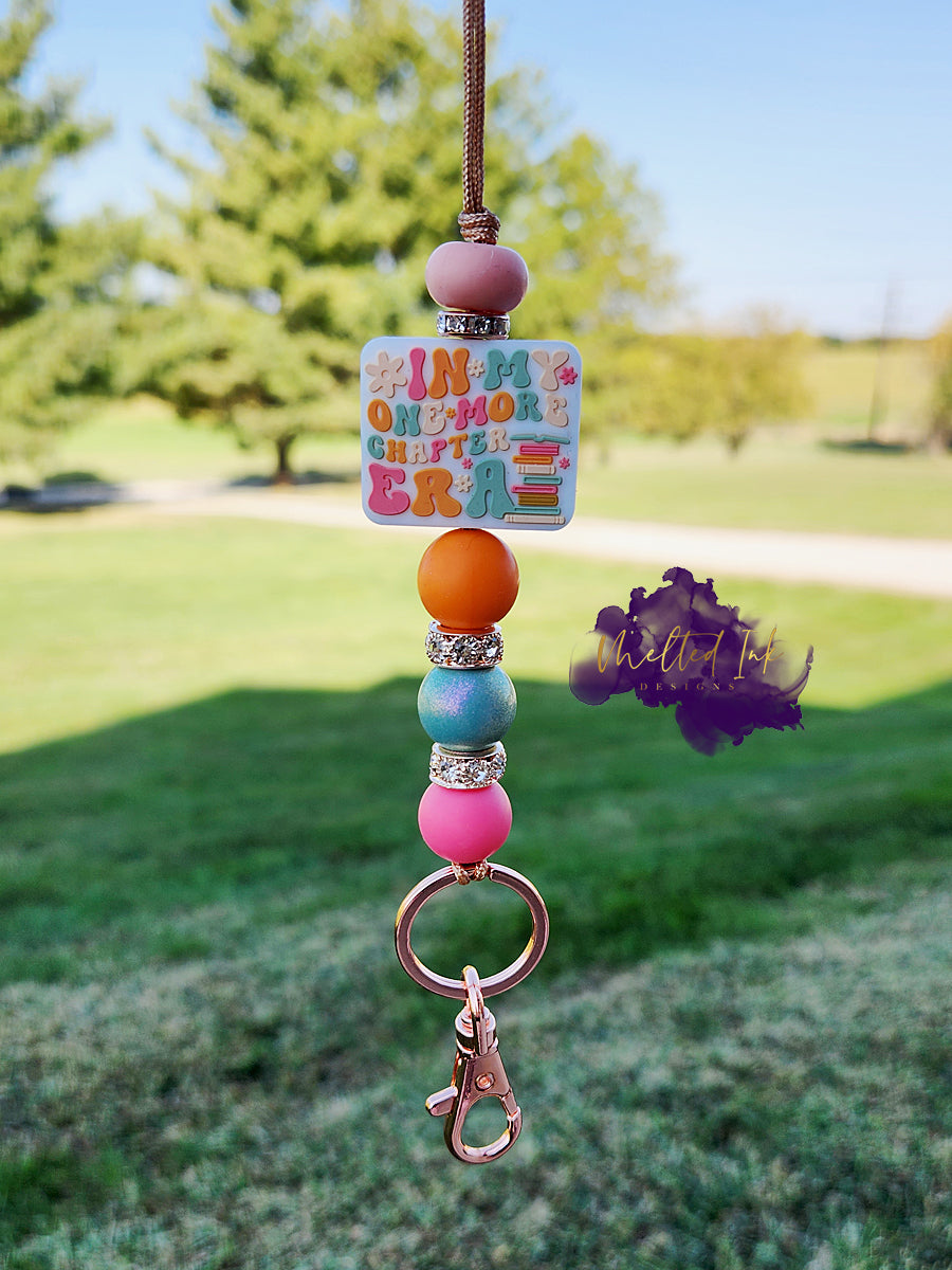 This lanyard features a focal bead that says "In My One More Chapter Era," adorned with cute little book illustrations. The design is accented with rhinestone spacers and colorful silicone beads in orange, bluish, and pink tones, creating a vibrant and playful look. It also includes a keyring for convenience. 