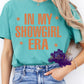 Shirt design featuring large block lettering that reads “In My Showgirl Era” with stars on each side of the text. The design uses warm orange text for bold contrast and a confident, retro vibe.