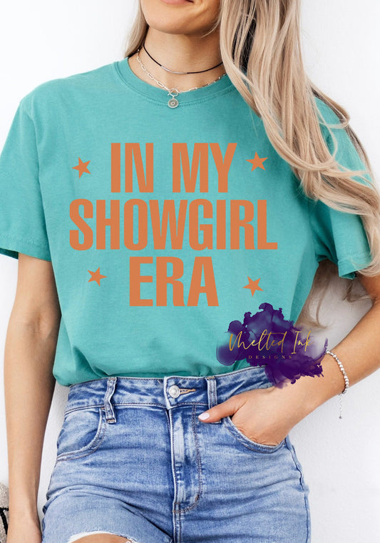Shirt design featuring large block lettering that reads “In My Showgirl Era” with stars on each side of the text. The design uses warm orange text for bold contrast and a confident, retro vibe.