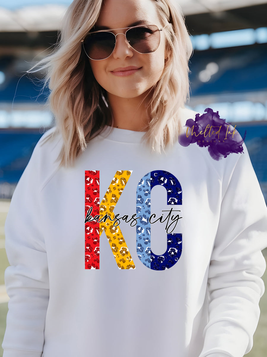 Photo is a mock up of design. Design is of the letters K and C. The K is red and yellow for KC Chiefs colors and the C is light and dark blue for the KC Royals. The letters are a faux glitter leopard print and across the letters the design says Kansas City in black. 