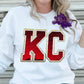 Photo is a mock up of design on a white sweatshirt. Design is of the letters KC with red sequins outlined with white sequins with a trim of gold. 