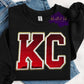 Photo is a mock up of design on a black sweatshirt. Design is of the letters KC with red sequins outlined with white sequins with a trim of gold. 