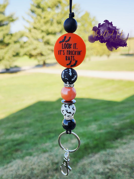 This Halloween lanyard features a fun focal bead that reads "Look, It's Frickin Bats" on one side and "I Love Halloween" on the other. It includes four 15mm silicone beads: a pearl black bead, a pearl orange bead, and two bat-themed beads—one white with black bats and the other black with white bats. The design is enhanced with rhinestone spacers, adding a touch of sparkle, and it comes with a keyring for practicality.