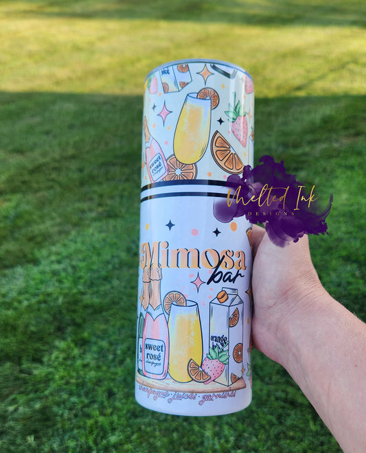 The photo showcases a vibrant 20oz tumbler that prominently displays the phrase "Mimosa Bar." The design is accented with playful elements of various alcohol drinks, along with colorful fruit slices, such as oranges, and strawberries, capturing the refreshing essence of mimosas.